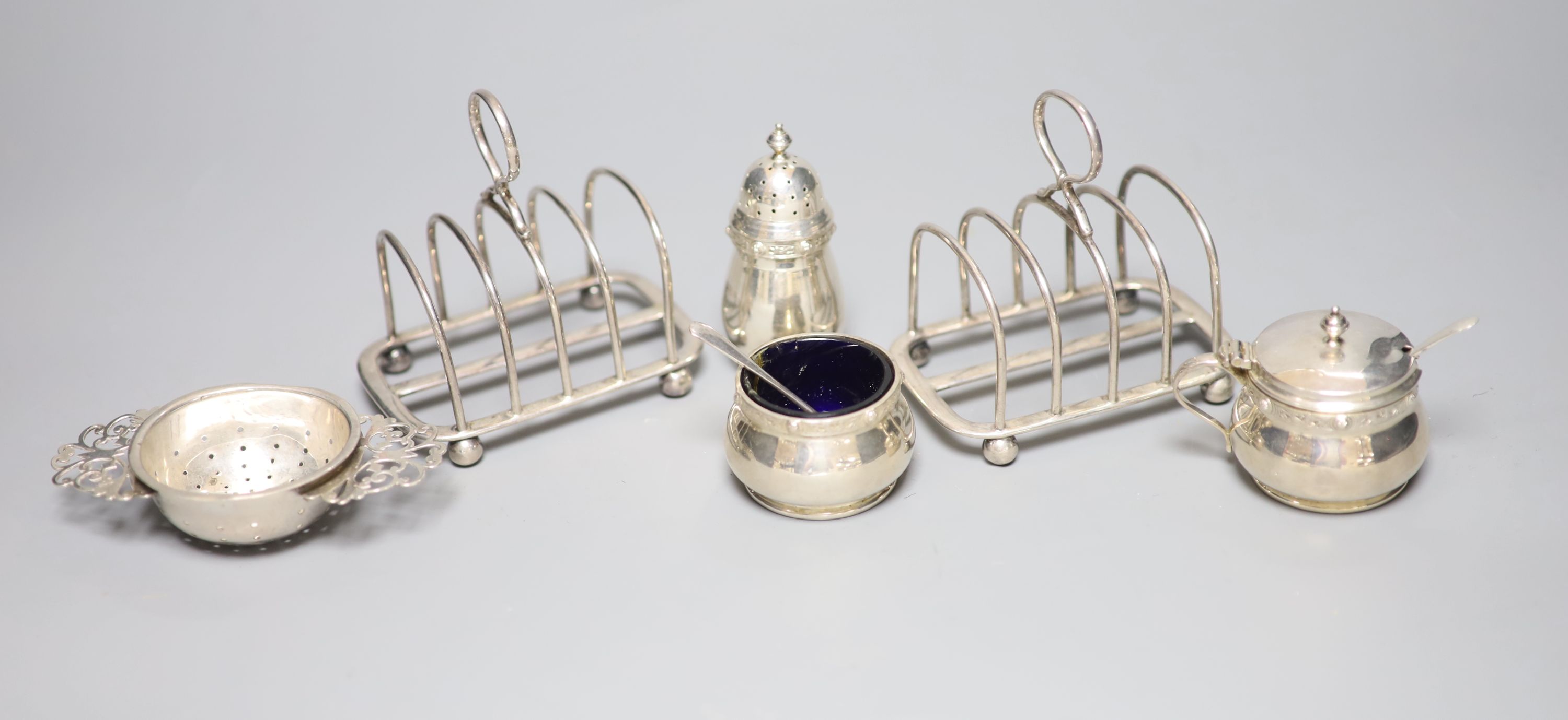 A pair of George V silver toast racks, a silver tea strainer and a silver three piece condiment set.